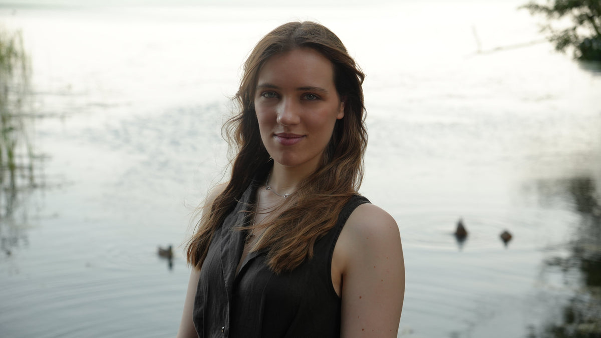 A woman, Julia Woodward, with long brown hair stands outdoors by a calm lake, wearing a sleeveless dark green dress. Ducks are visible in the water behind her, and the background is softly blurred.